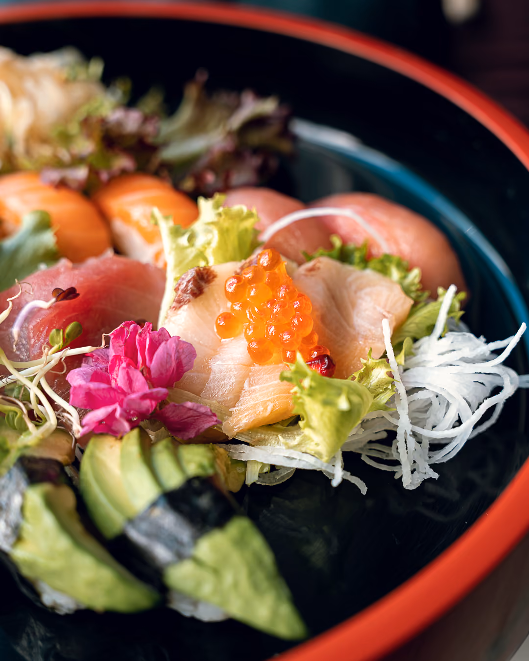 Detailaufnahme einer Sushi-Platte mit Sashimi, Lachsrogen, Avocado und einer rosa Essblüte in roter Lackschale.