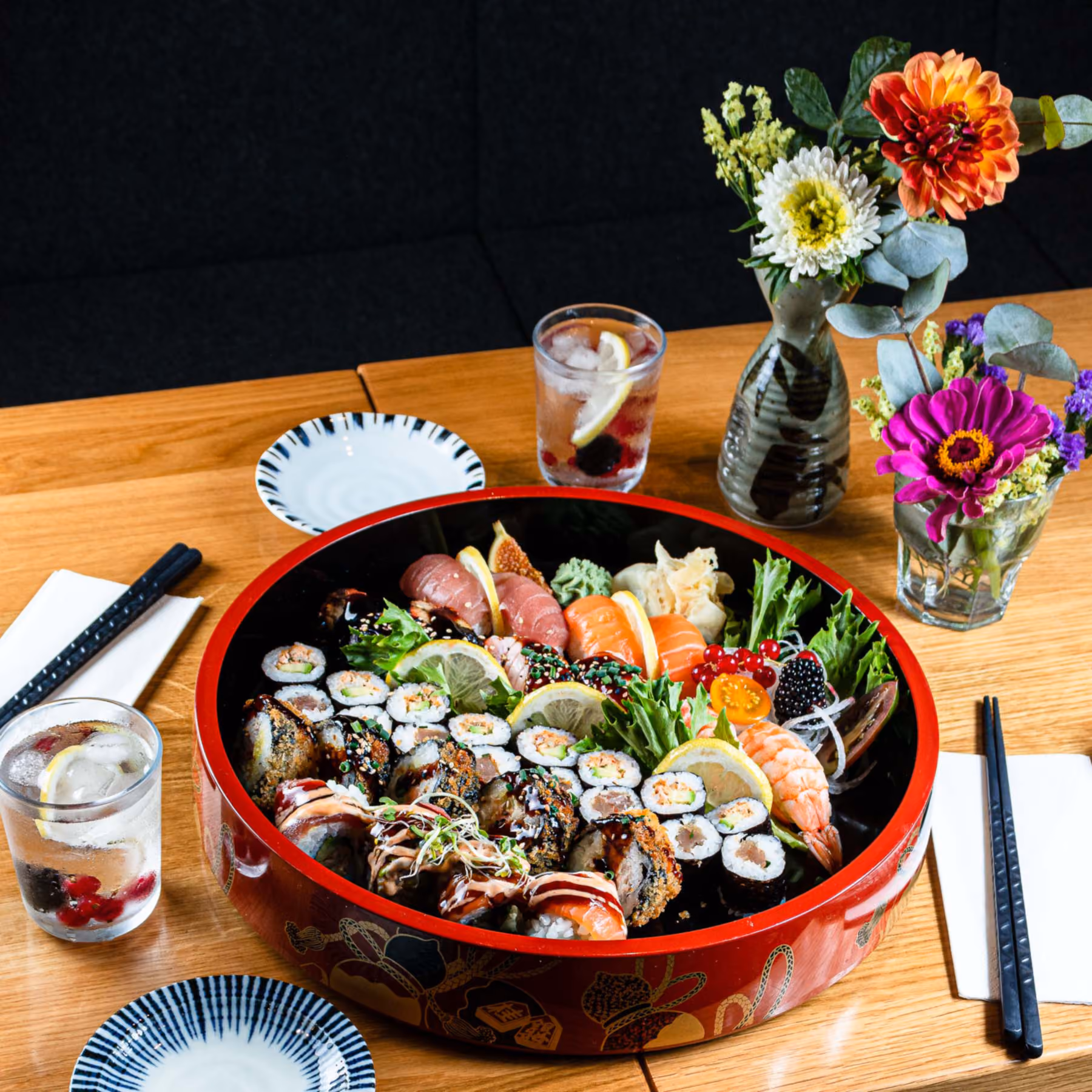 Große rote Lackschale voller Maki, Nigiri und Garnelen auf einem Holztisch, daneben Getränke, Stäbchen und Blumenvasen.